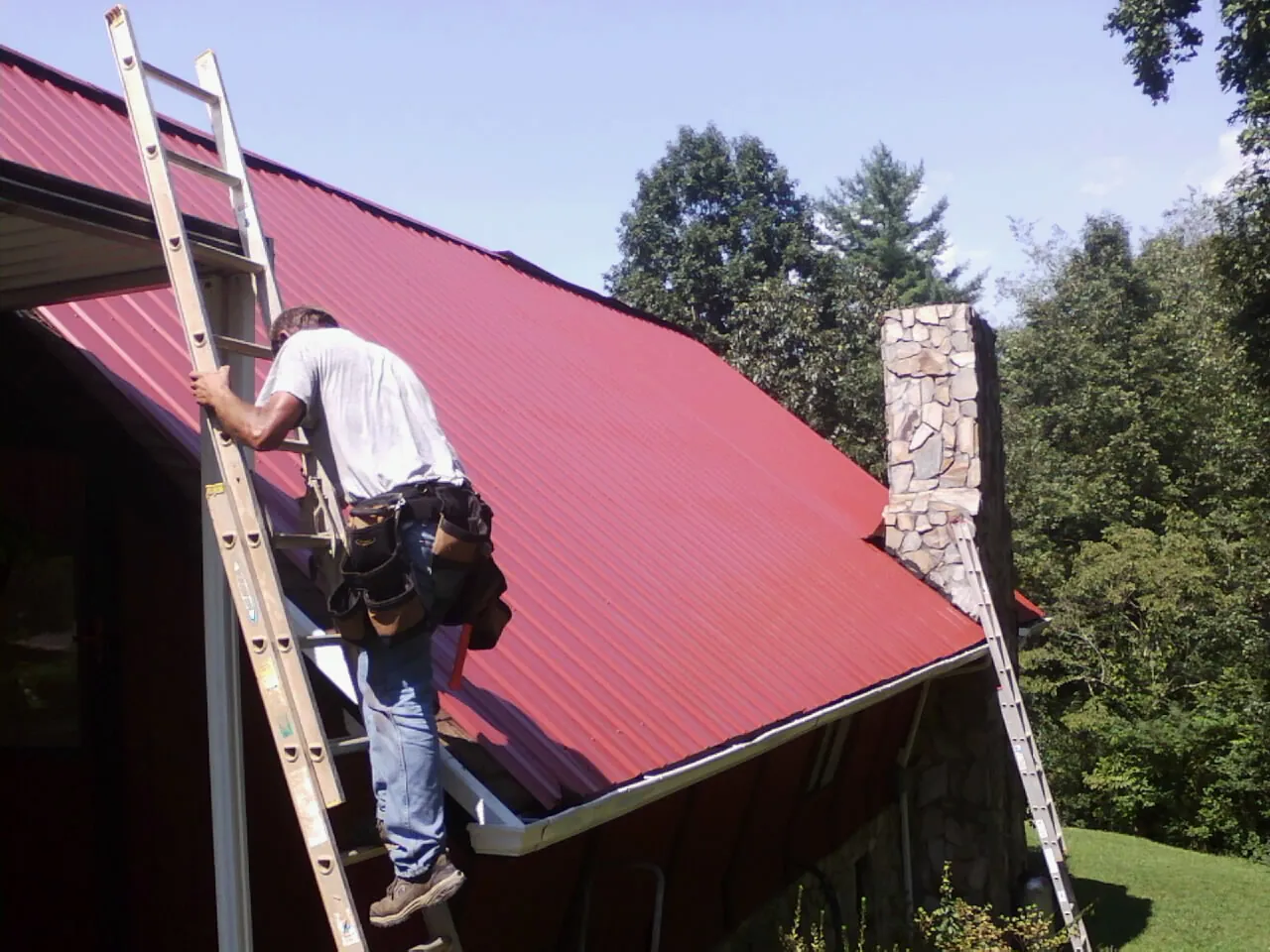 Roofer on a ladder beside a red metal roof and stone chimney — Soffit & Fascia Repair by Roofing Brothers in Muncy